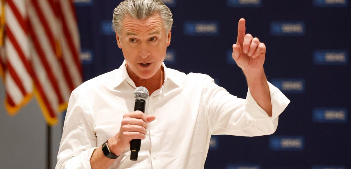 Gavin Newsom A man in a white button shirt holding a microphone with an American flag in the background