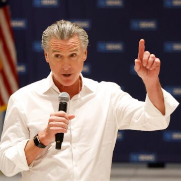 Gavin Newsom Un hombre con una camisa blanca abotonada sostiene un micrófono con una bandera estadounidense de fondo.