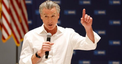 A man in a white button shirt holding a microphone with an American flag in the background