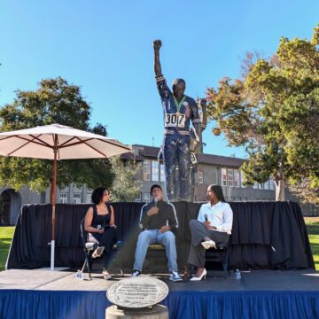 John Carlos -SJSU-2 resized Men sitting on a chair below his bronze statute