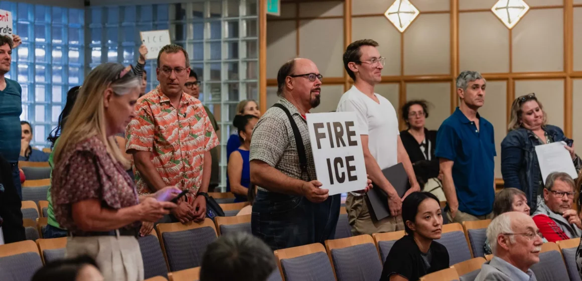 People standing at a city council meeting, with some holding protest signs, in Mountain View, California