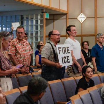 People standing at a city council meeting, with some holding protest signs, in Mountain View, California