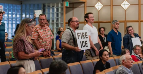 People standing at a city council meeting, with some holding protest signs, in Mountain View, California