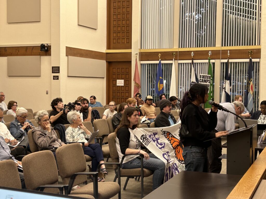 People sitting in chairs at a government meeting in Gilroy, California, with one speaker standing at the podium