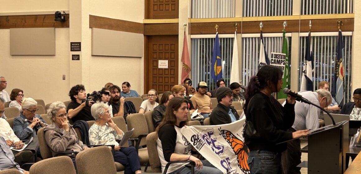 People sitting in chairs at a government meeting in Gilroy, California, with one speaker standing at the podium