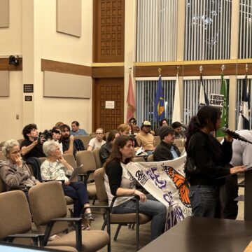 People sitting in chairs at a government meeting in Gilroy, California, with one speaker standing at the podium