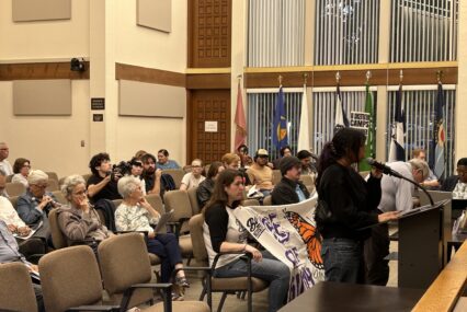 People sitting in chairs at a government meeting in Gilroy, California, with one speaker standing at the podium