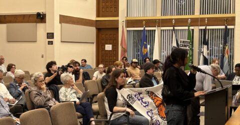 People sitting in chairs at a government meeting in Gilroy, California, with one speaker standing at the podium