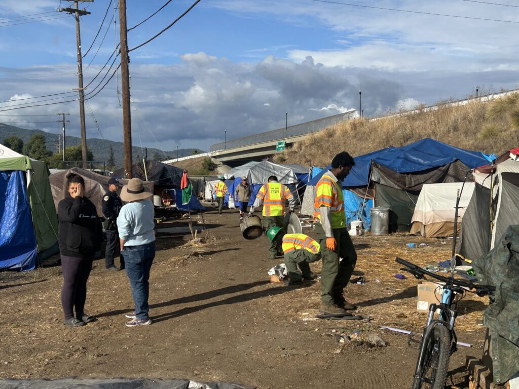 People standing around a homeless camp while workers cleanup nearby trash and debris in Gilroy, California
