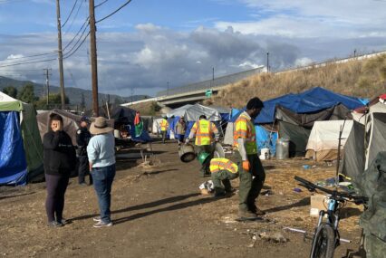 People standing around a homeless camp while workers cleanup nearby trash and debris in Gilroy, California