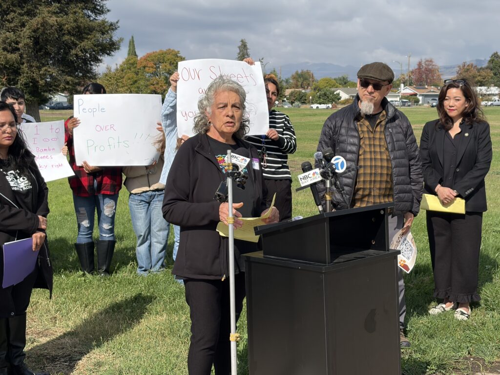 A woman standing outside at a podium, with people behind her holding protest signs