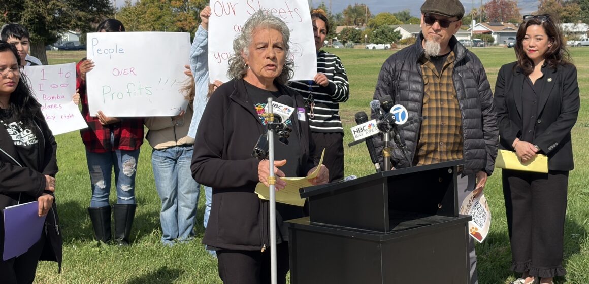 A woman standing outside at a podium, with people behind her holding protest signs