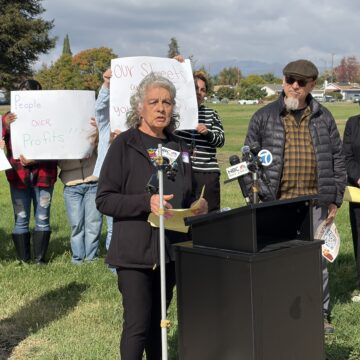 A woman standing outside at a podium, with people behind her holding protest signs