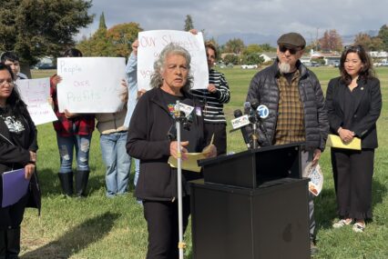 A woman standing outside at a podium, with people behind her holding protest signs
