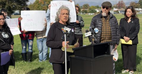 A woman standing outside at a podium, with people behind her holding protest signs